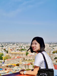 A photo of Mianmian looking over her shoulder and smiling. She is leaning on a railing and overseeing a cityscape in the distance.